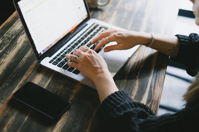 A person typing on a laptop beside a cell phone